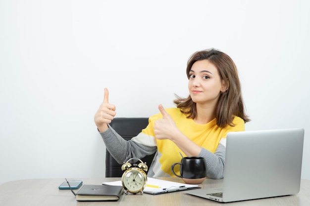 Photo of female office worker sitting at the desk and showing thumbs up . High quality photo