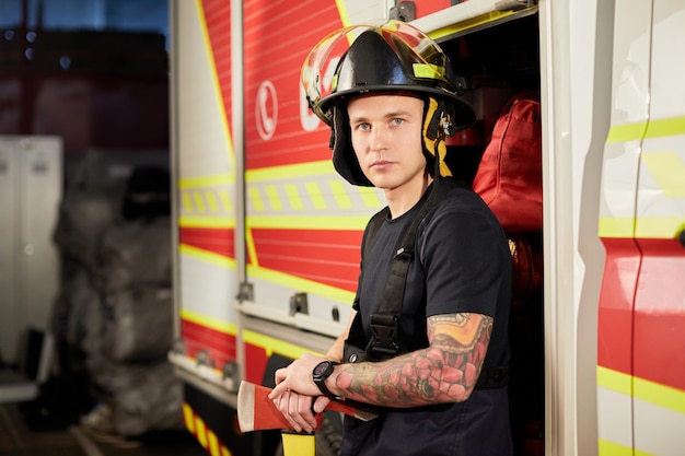 Photo of fireman wearing helmet with ax against fire engine Image of young man firefighter with helmet in head against background of fire truck