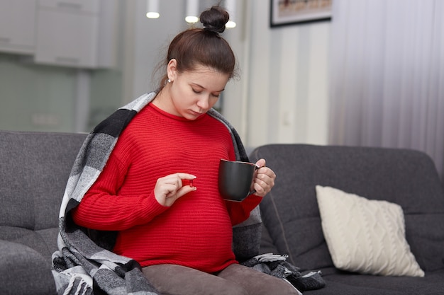 Photo fo pregnant female takes medicine, holds cup and pill, needs vitamins, dressed casually, sits at couch