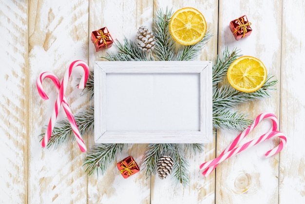 Photo frame between Christmas decoration, with candy canes and gift boxs on a white wooden table. Top view, frame to copy space