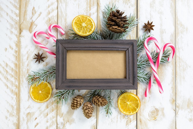 Photo frame between Christmas decoration, with candy canes and pine cones on a white wooden table. Top view, frame to copy space