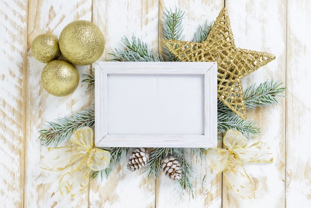 Photo frame between Christmas decoration, with golden color balls and star on a white wooden table. Top view, frame to copy space