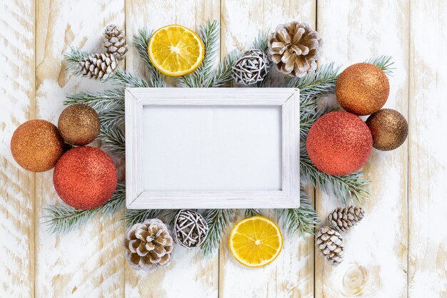 Photo frame between Christmas decoration, with multicolored balls and pine cones on a white wooden table. Top view, frame to copy space