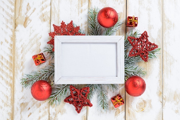 Photo frame between Christmas decoration, with red balls and stars on a white wooden table. Top view, frame to copy space