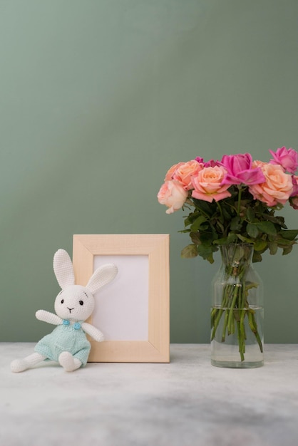 녹색 배경에 장미 꽃병과 니트 장난감 토끼가 있는 텍스트용 사진 프레임 모의