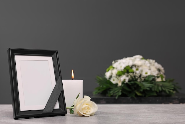 photo frame with black ribbon burning candle rose on light table and wreath of flowers near grey wall indoors space for text funeral attributes