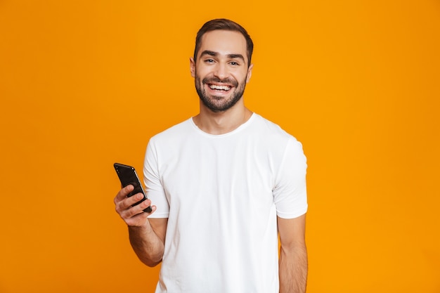 Photo of friendly man 30s in casual wear smiling and holding smartphone, isolated