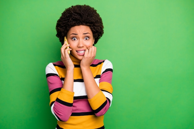 Photo of frightened dark skin wavy lady hold telephone speak best friend terrified listening scary story bite fingers wear casual striped jumper jeans 