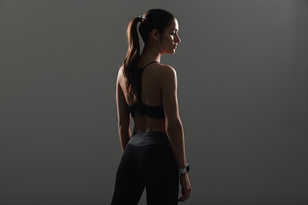 Photo from back of beautiful fitness woman posing  in sportwear with stopwatch on wrist, isolated over dark wall