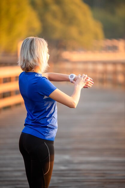 La foto dalla parte posteriore della donna sportiva guarda l'orologio a portata di mano, in piedi sul ponte di legno nel parco il giorno d'estate