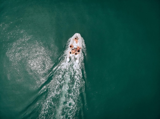 Photo from a bird's eye view. Girls and a guy ride a white boat in the open sea on a hot day. The guys are enjoying the ride. The guy in the cap sits at the wheel. Admiration for girls.