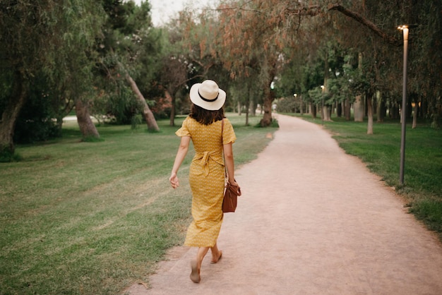 A photo from behind of a brunette girl in a yellow dress and a hat who is walking on a sand path in the Valencian park in the evening.