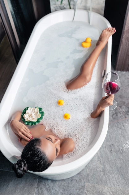 Photo from above: Girl posing while lying in a foam bath, with a flower and ducks, with a glass of red wine in her hand, enjoying relaxation. Enjoyment. Personal care.
