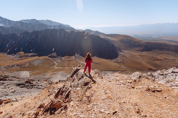 A photo from a high angle with a young girl in a red suit standing with her back and looking at the rocky mountains