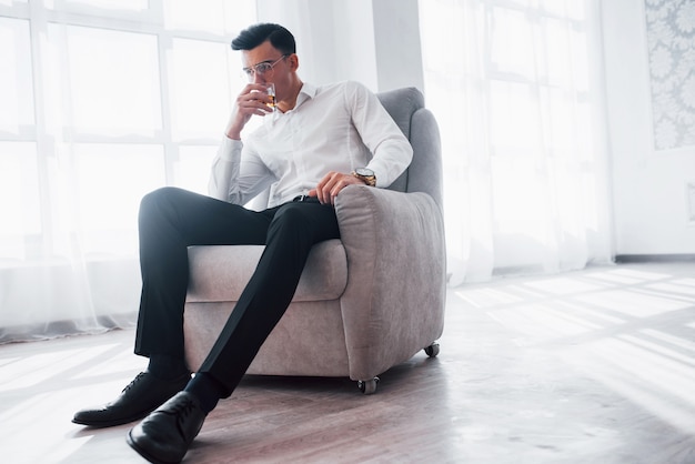 Photo from below. Luxury looking man in the classic wear sitting on the chair in the room and holding the glass with alcohol.