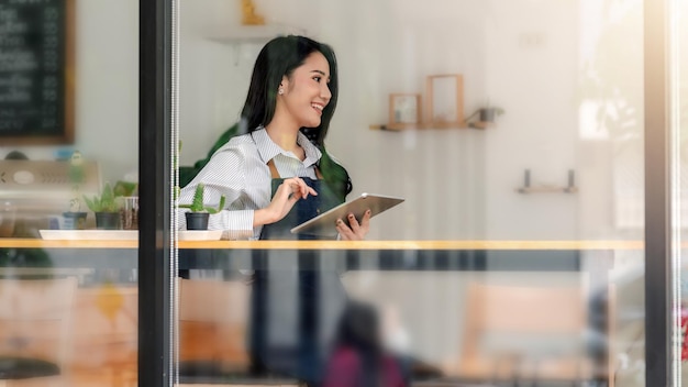 카페에서 태블릿을 들고 카운터에 앉아 있는 행복한 아시아 여성의 작은 비즈니스 소유자의 유리 측면 보기 외부에서 사진.