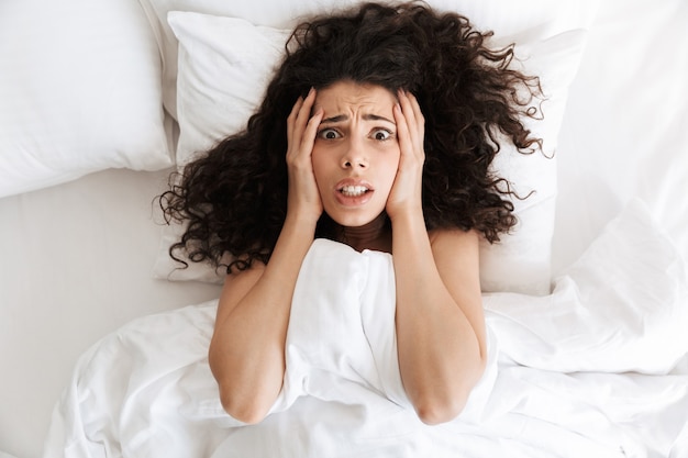 Foto foto dall'alto di una donna in preda al panico 20s con i capelli ricci scuri che afferra la testa dopo un brutto sogno, mentre giaceva a letto con una coperta bianca