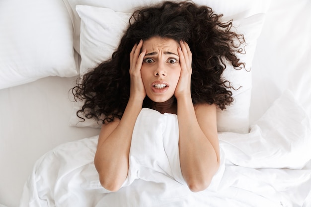 Photo from above of panic woman 20s with dark curly hair grabbing head after bad dream, while lying in bed with white blanket