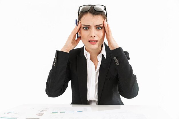 Foto foto di una donna d'affari frustrata lavoratrice vestita in abiti formali che si tocca la testa mentre lavora e si siede alla scrivania in ufficio isolata su un muro bianco