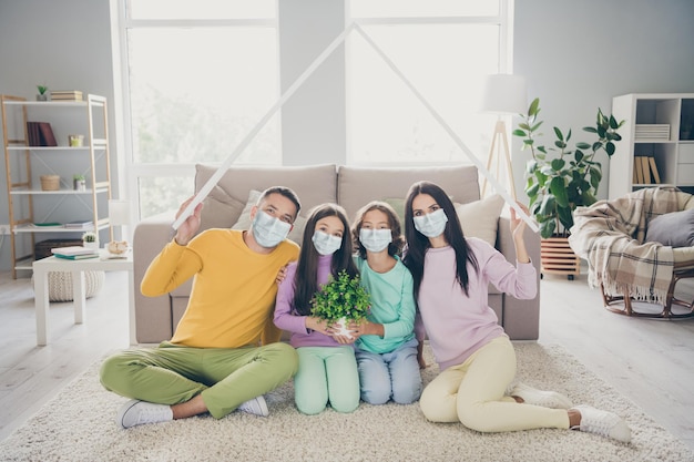 Photo of full family four people sit carpet dad mom hold paper roof children flower wear mask colorful jumper pants in living room indoors