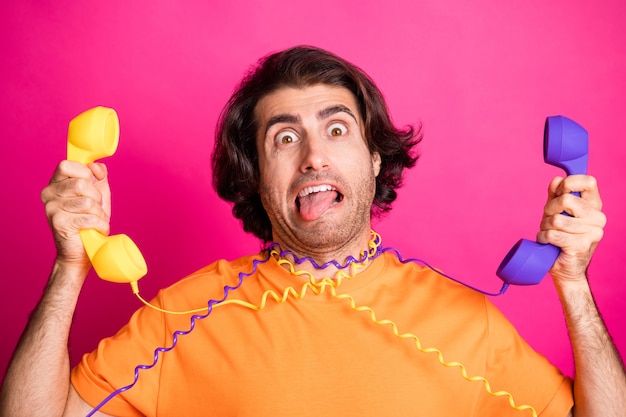 Photo photo of funky guy tongue-out arms hold two telephones hang cables wear orange t-shirt isolated pink color background