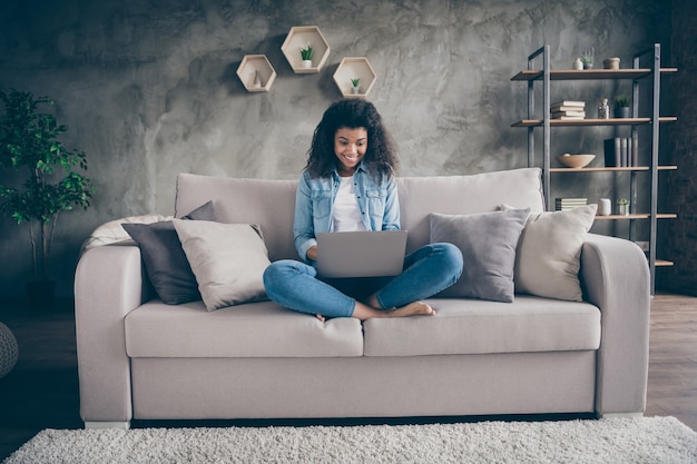 Photo of funny amazed dark skin wavy lady notebook on knees crossed legs texting friends read good news sitting cozy couch casual denim outfit living room indoors