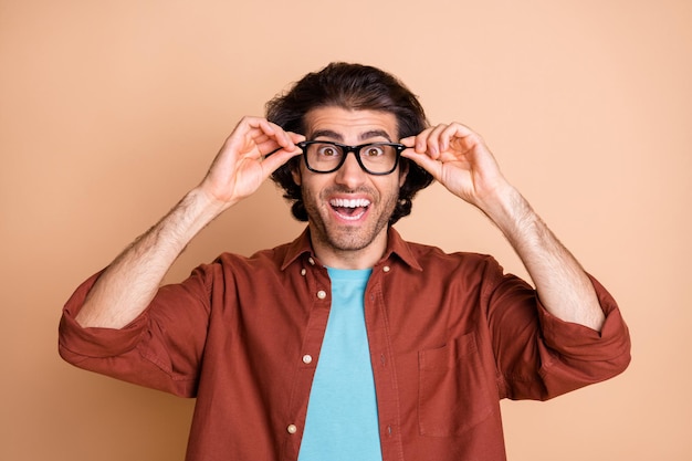 Photo of funny cheerful guy arms hold eyewear crazy face wear glasses brown t-shirt isolated beige color background