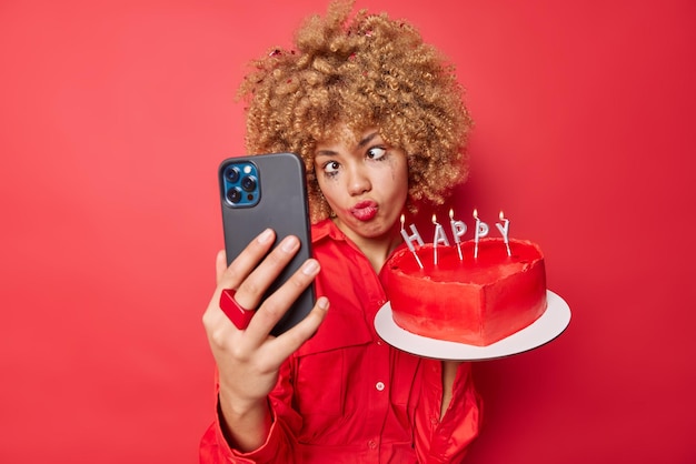 Photo of funny curly haired European woman crosses eyes keeps lips folded takes selfie via smartphone has leaked makeup wears shirt isolated over red background holds heart cake with candles