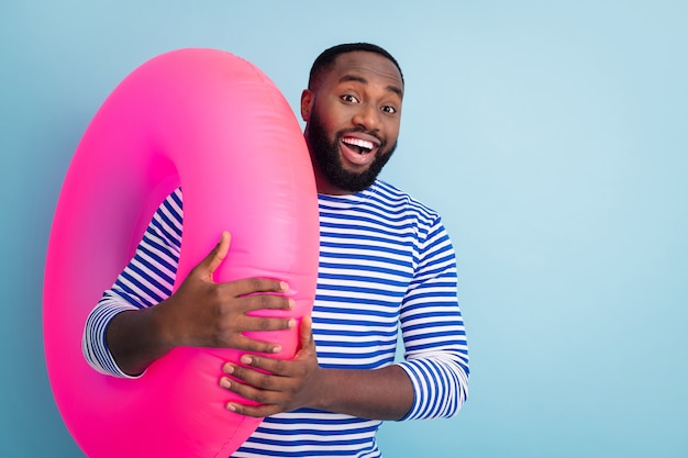 Photo of funny dark skin guy holding pink life buoy ready to swim ocean sea addicted traveler good mood sunny day wear striped sailor shirt isolated blue color wall