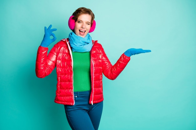 Photo of funny lady hold open arm novelty product cool offer show okay symbol wink eye wear casual red overcoat blue scarf pink ear covers jumper pants isolated teal color wall