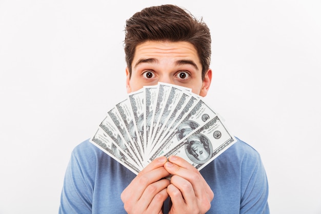 흰 벽 위에 고립 된 달러 통화에 돈의 팬으로 그의 얼굴을 덮고 캐주얼 티셔츠에 재미있는 사람의 사진