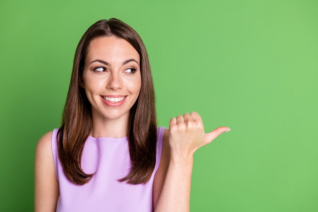 Photo girlish adorable lady model toothy smile point thumb look empty space demonstrate interesting product low price reliable service dressed violet tank-top isolated green color background