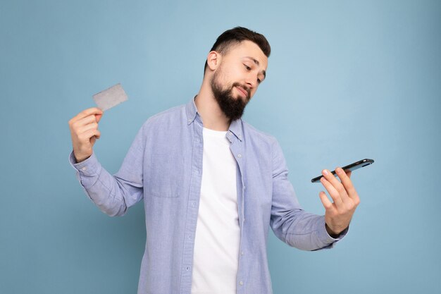 Photo of good looking attractive brunet unshaven young man wearing casual blue shirt and white t-shirt isolated over blue background wall holding credit card and using mobile phone making payment look