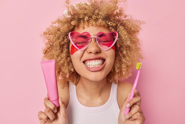 Photo of good looking curly woman shows white well cared teeth\
after brushing holds tube of toothpaste and toothbrush applies\
beauty patches under eyes wears sunglasses and t shirt poses\
indoor