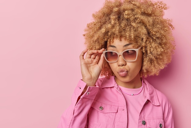 Photo of good looking young european woman with curly hair\
keeps lips folded wears sunglasses and jacket has stylish look\
isolated over pink background blank space for your advertising\
content