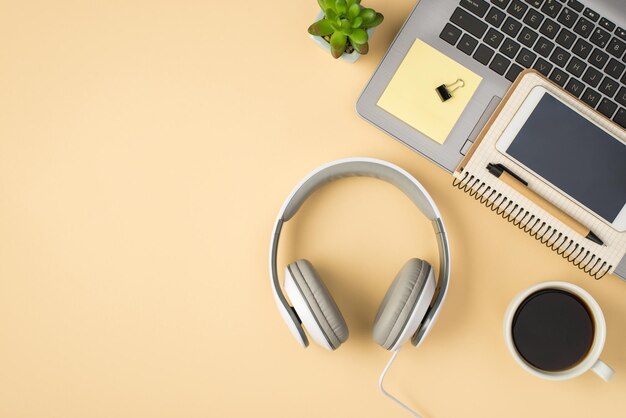 Above photo of grey laptop cup of coffee notepad with pen phone paperclips headphones plant and stickers isolated on the beige backdrop with blank space