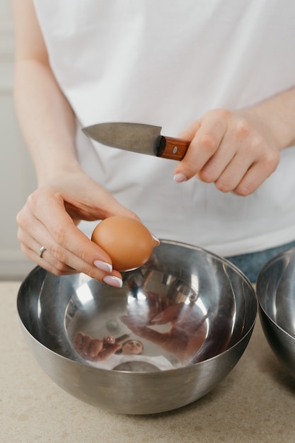 A photo of the hands of a young woman who is breaking the eggshell with the knife above the stainless steel soup bowl in the kitchen