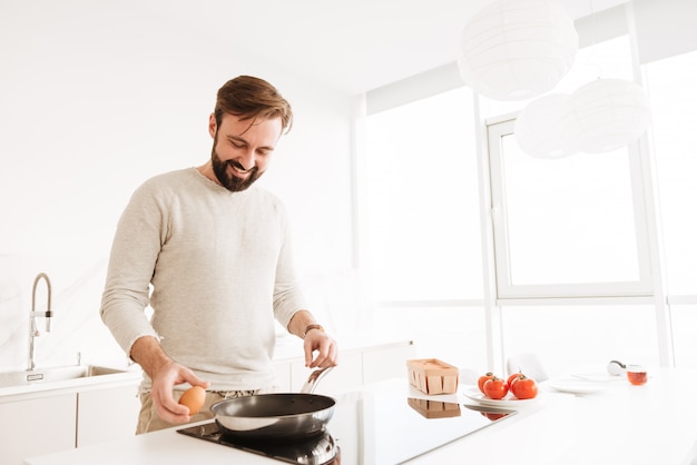 Фото красивого холостяка с короткими каштановыми волосами и бородой, готовим омлет с овощами в домашней кухне, используя сковороду