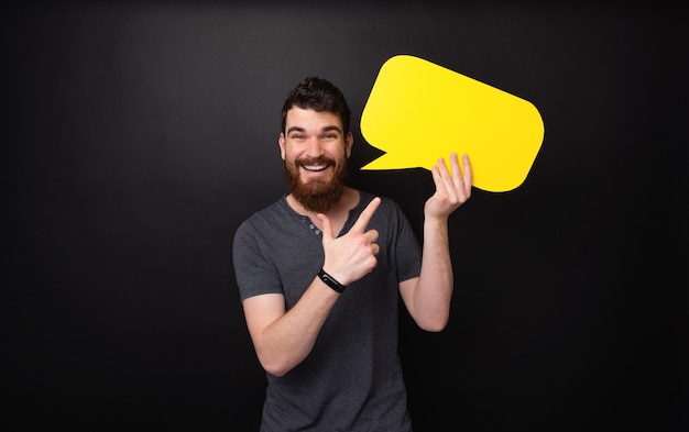 Photo of handsome bearded guy pointing at yellow bubble speech over dark background