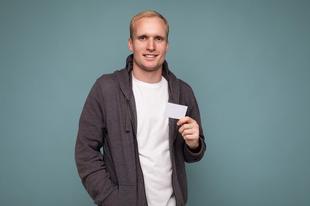 Photo of handsome blonde male person wearing grey sweater and white t-shirt isolated over blue background wall holding credit card looking at camera.