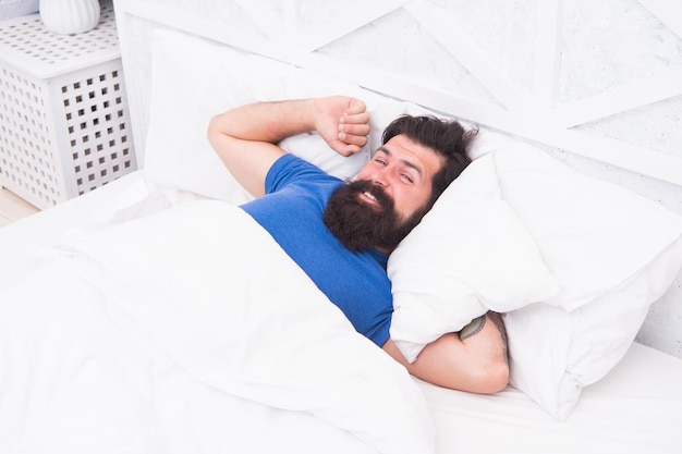 Photo of handsome cheerful woke up man lies in bed indoors at home looking camera relax and recreation