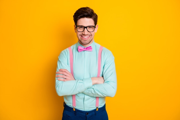 Фото красивой крутой одежды деловой парень стоит самоуверенно скрещенными руками сияющая улыбка носить спецификации рубашка подтяжки галстук-бабочка брюки изолированные желтый цвет фона