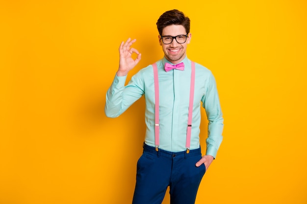 Photo of handsome cool clothes guy boyfriend selfconfident\
person showing okey symbol express agreement wear specs shirt\
suspenders bow tie trousers isolated yellow color background