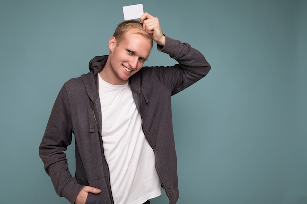 Photo of handsome happy smiling blonde man wearing grey sweater and white t-shirt isolated over blue background wall holding credit card looking at camera. copy space