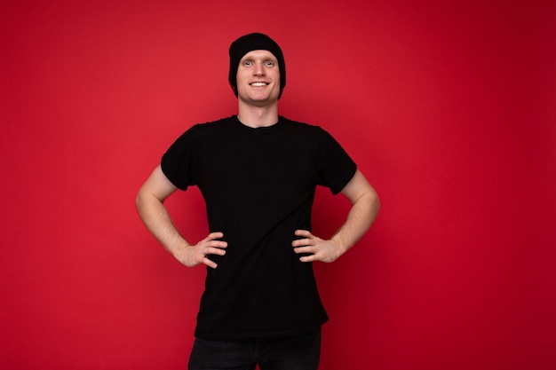 Photo of handsome happy smiling young man standing isolated over red background wall wearing black t-shirt for mockup and black hat and looking at camera.