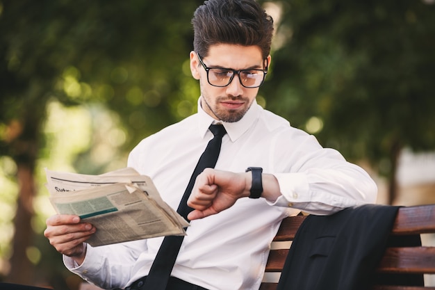 Photo of handsome man in businesslike suit reading newspaper on bench in green park, and looking at wristwatch