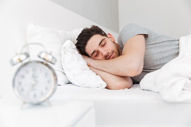 Foto foto di bell'uomo con stoppie e indossa abiti casual, dormendo a casa a letto con l'orologio sul comodino