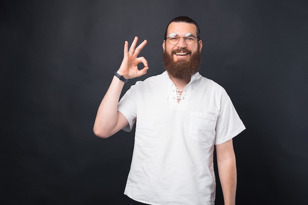 Photo of handsome man with beard in white t-shirt showing ok gesture over black background