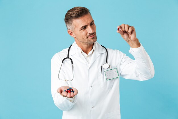 Photo of a handsome mature man doctor posing isolated over blue wall wall holding pills vitamins.