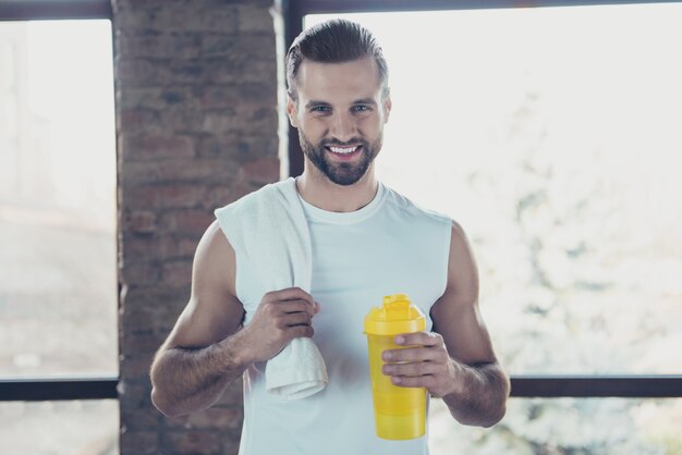 Photo of handsome sportsman trainer guy wait morning practicing hold water bottle towel friendly smiling sportswear tank-top training house  windows indoors